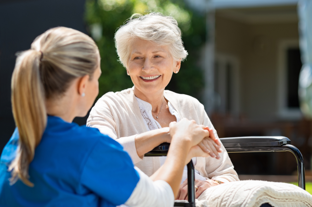 Smiling,Senior,Patient,Sitting,On,Wheelchair,With,Nurse,Supporting,Her.